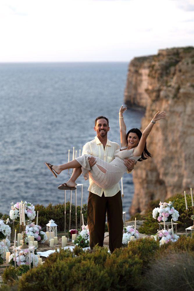 A man lifting his new fiancee in his arms, both laughing, on a clifftop lined with flowers