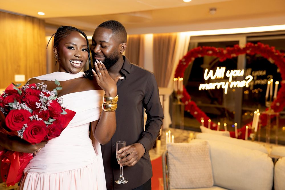 A newly engaged couple indoors with a red rose bouquet, the neon sign glowing behind them