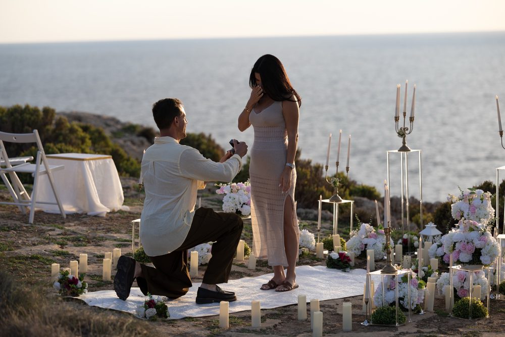 A man on one knee on the clifftop as she brings her hand to her face