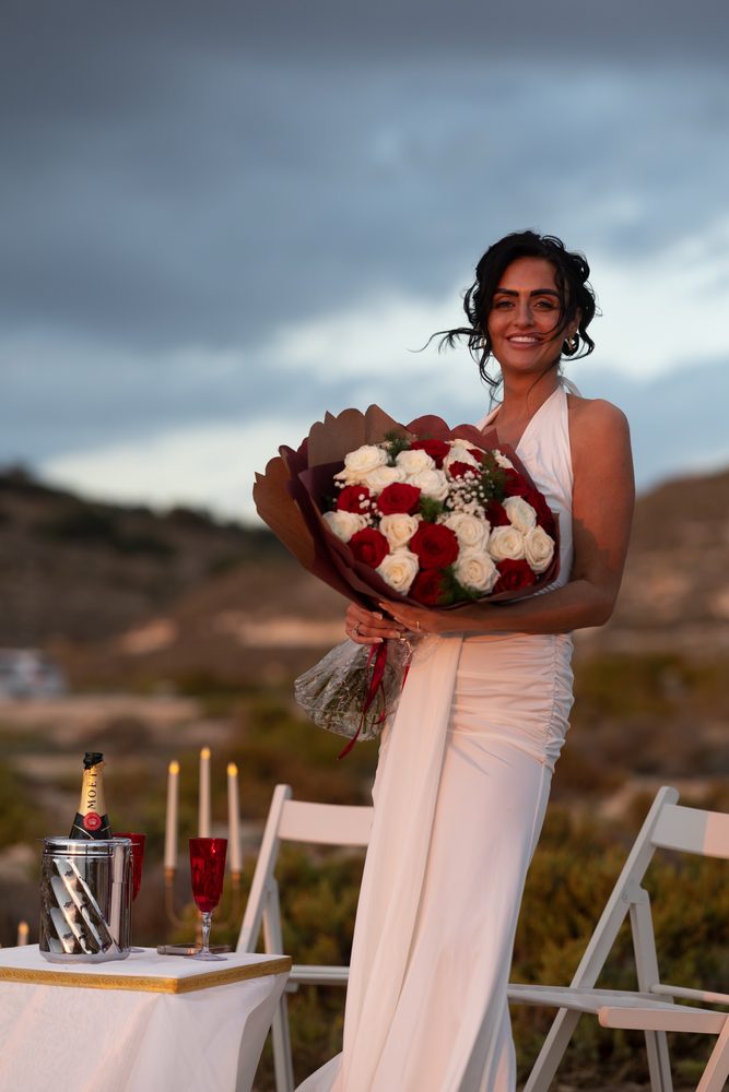 A woman holding a red and white rose bouquet with the sea behind her