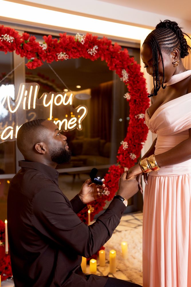 A man on one knee holding the ring box, the red heart arch behind him